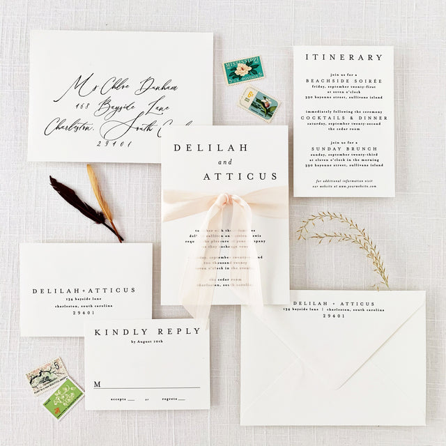 A set of white wedding invitations with minimalist design, including an envelope and RSVP card, displayed on a table with a silk ribbon and stylized text.