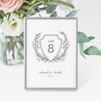 A grey and white wedding table number card featuring a traditional laurel crest design with a space for the table number and names at the bottom.
