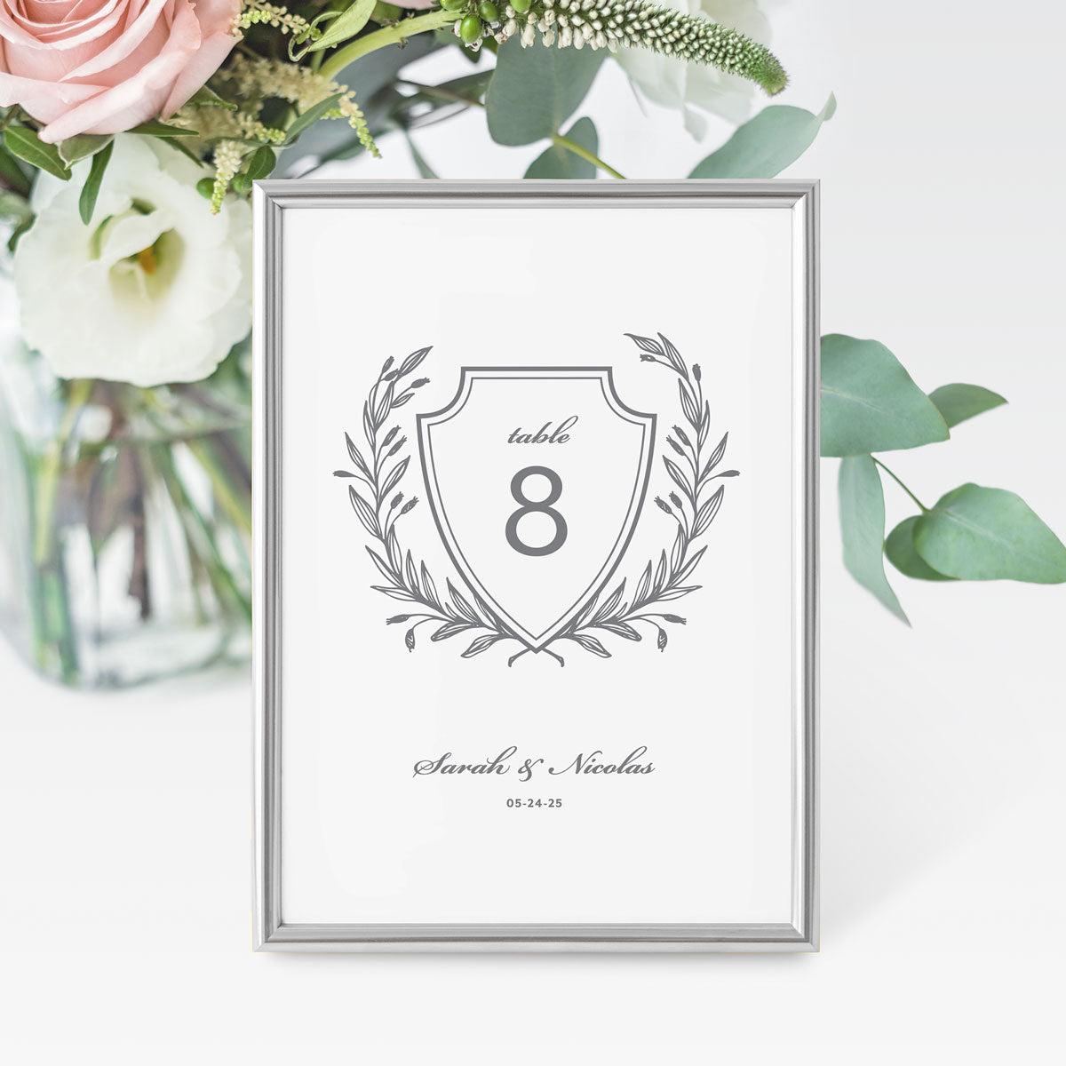 A grey and white wedding table number card featuring a traditional laurel crest design with a space for the table number and names at the bottom.