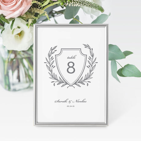 A grey and white wedding table number card featuring a traditional laurel crest design with a space for the table number and names at the bottom.