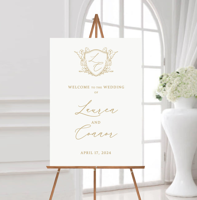 white and gold wedding welcome sign featuring a monogrammed crest at the top and the names of the couple in flowing calligraphy script fonts