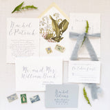A set of dusty blue script wedding invitations with envelope, RSVP card, and decorative elements like flowers and a postage stamp on a white background.