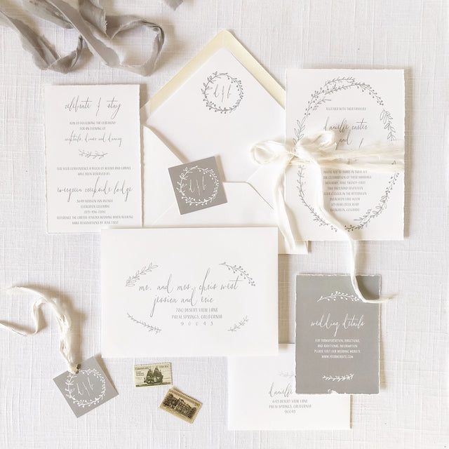 A set of white wedding invitations with hand-torn edges and a minimal wreath design, accompanied by envelopes and RSVP cards, displayed on a linen surface with a grey ribbon.